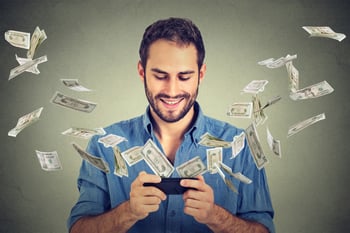 Technology online banking money transfer, e-commerce concept. Happy young man using smartphone with dollar bills flying away from screen isolated on gray wall office background.